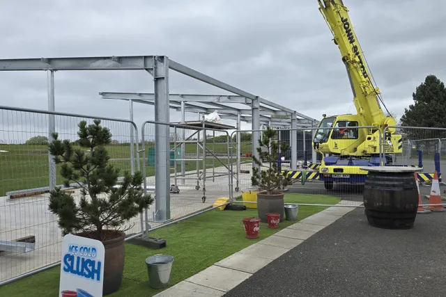 Steel canopy frame under construction alongside the crane, driving range visible beyond the site fencing
